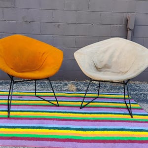 May include: Two diamond-shaped lounge chairs with thin black metal wire frames. One chair features a vibrant mustard yellow fabric upholstery, while the other is covered in a neutral off-white fabric. Both chairs have a wide, low-profile seat.