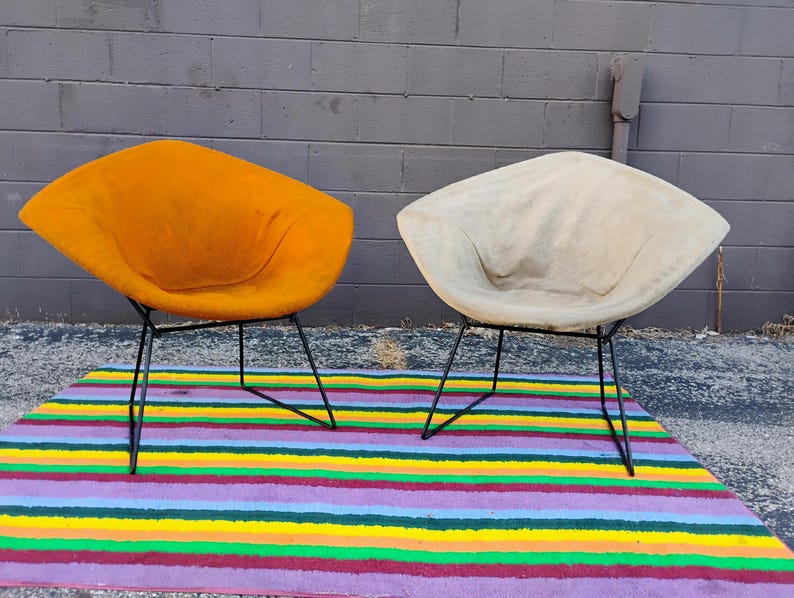 May include: Two diamond-shaped lounge chairs with thin black metal wire frames. One chair features a vibrant mustard yellow fabric upholstery, while the other is covered in a neutral off-white fabric. Both chairs have a wide, low-profile seat.