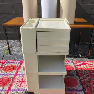May include: A vintage, off-white rolling storage cart with drawers and open shelves. The cart is flanked by three cylindrical planters and two small wooden tables with metal legs. The background is a dark gray brick wall.