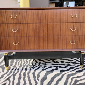 May include: A vintage wooden dresser with three drawers and black legs. The drawers have brass handles and the legs have gold accents.