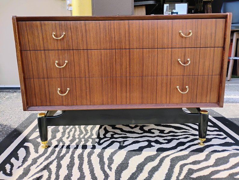 May include: A vintage wooden dresser with three drawers and black legs. The drawers have brass handles and the legs have gold accents.