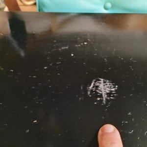 May include: Close-up of a black, glossy surface with multiple small white specks and a cluster of scratches. A finger points towards the scratched area. The surface appears to be part of a larger object.