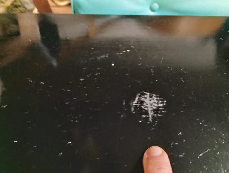 May include: Close-up of a black, glossy surface with multiple small white specks and a cluster of scratches. A finger points towards the scratched area. The surface appears to be part of a larger object.