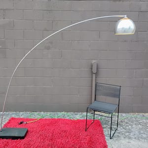 K&ouml;nnte beinhalten: Eine moderne Bogen-Stehleuchte mit silbernem Kuppel-Schirm und wei&szlig;em, gebogenem Arm. Die Lampe steht auf einem roten Shaggy-Teppich mit schwarzem Sockel. Ein schwarzer Metallstuhl steht neben dem Teppich.
