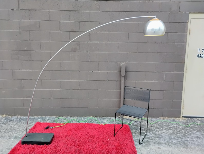 K&ouml;nnte beinhalten: Eine moderne Bogen-Stehleuchte mit silbernem Kuppel-Schirm und wei&szlig;em, gebogenem Arm. Die Lampe steht auf einem roten Shaggy-Teppich mit schwarzem Sockel. Ein schwarzer Metallstuhl steht neben dem Teppich.