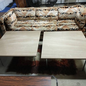 May include: A vintage three-seat sofa with a geometric pattern in shades of brown and beige. Two square, light-colored wooden coffee tables with metal legs are in front of the sofa. A shag rug and other furniture are in the background.