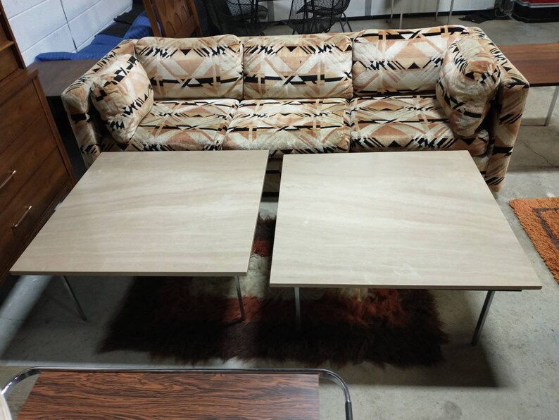 May include: A vintage three-seat sofa with a geometric pattern in shades of brown and beige. Two square, light-colored wooden coffee tables with metal legs are in front of the sofa. A shag rug and other furniture are in the background.