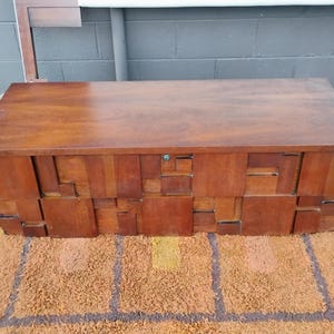 Puede incluir: Un cofre de madera rectangular con un acabado marr&oacute;n oscuro. El cofre tiene una parte superior plana y una parte frontal con un patr&oacute;n geom&eacute;trico de bloques rectangulares. El cofre est&aacute; sobre una alfombra estampada marr&oacute;n y naranja.