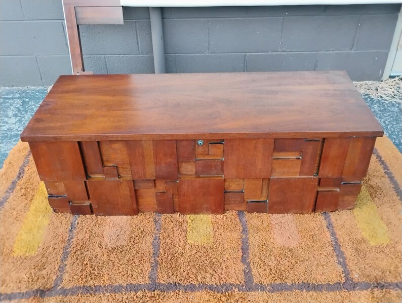 Puede incluir: Un cofre de madera rectangular con un acabado marr&oacute;n oscuro. El cofre tiene una parte superior plana y una parte frontal con un patr&oacute;n geom&eacute;trico de bloques rectangulares. El cofre est&aacute; sobre una alfombra estampada marr&oacute;n y naranja.
