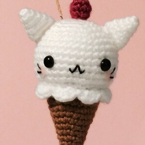 May include: A white crocheted ice cream cone with cat ears and a red cherry on top. The ice cream cone has black eyes and a black mouth.