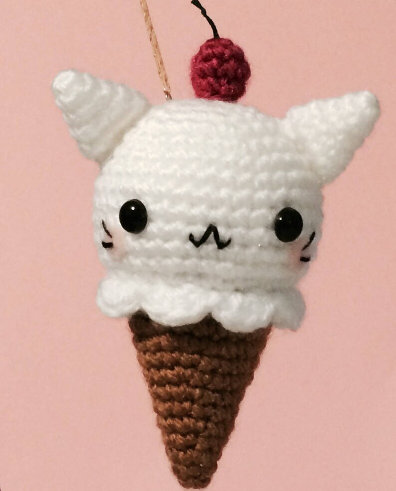 May include: A white crocheted ice cream cone with cat ears and a red cherry on top. The ice cream cone has black eyes and a black mouth.