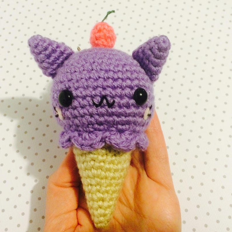 May include: Purple crocheted ice cream cone with a pink cherry on top and black eyes. The ice cream cone has a cute cat face.