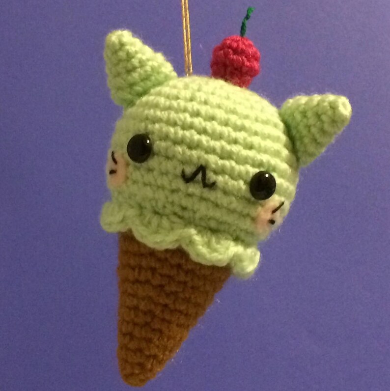 May include: A green crocheted ice cream cone with a cherry on top and cat ears. The cone has a cute cartoon face with black eyes and a pink nose.
