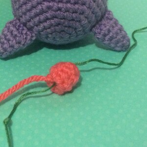 May include: A close-up of a pink crocheted ball with green and pink yarn attached to it. The ball is on a turquoise surface.