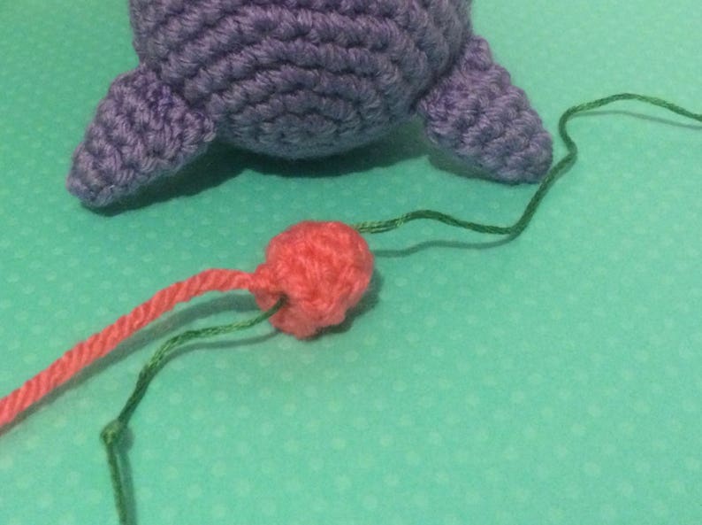 May include: A close-up of a pink crocheted ball with green and pink yarn attached to it. The ball is on a turquoise surface.