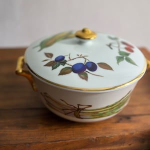 May include: A white porcelain covered dish with a gold trim and a floral design featuring plums and leaves. The dish has gold handles and sits on a wooden surface.