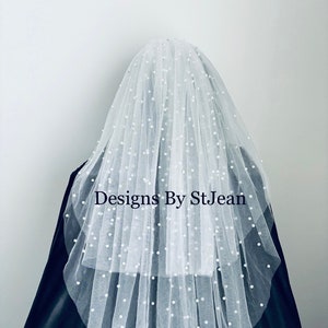 Puede incluir: Un velo de novia de tul blanco con adornos de perlas. El velo está en capas y tiene un borde festoneado. El texto "Designs By StJean" es visible en el centro del velo.