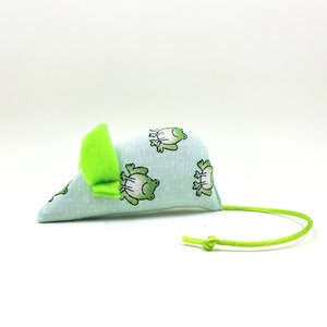 Puede incluir: Un juguete para gatos de color azul claro con forma de ratón, decorado con un estampado de ranas verdes. Tiene una oreja de fieltro verde brillante y una cola de cuerda verde. Juguete para gatos.