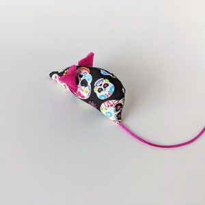May include: A black and white cat toy shaped like a mouse with a pink tail. The mouse is covered in a fabric with a pattern of colorful sugar skulls.
