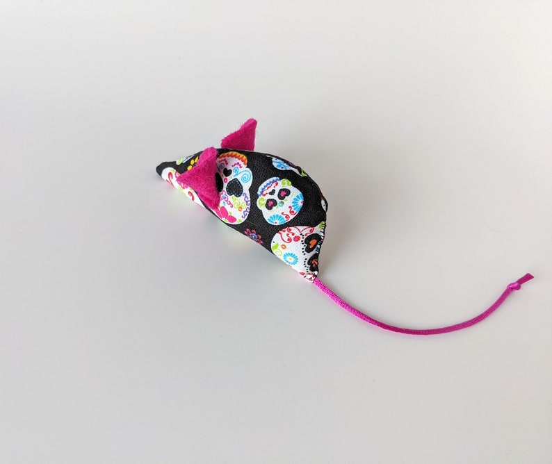 May include: A black and white cat toy shaped like a mouse with a pink tail. The mouse is covered in a fabric with a pattern of colorful sugar skulls.