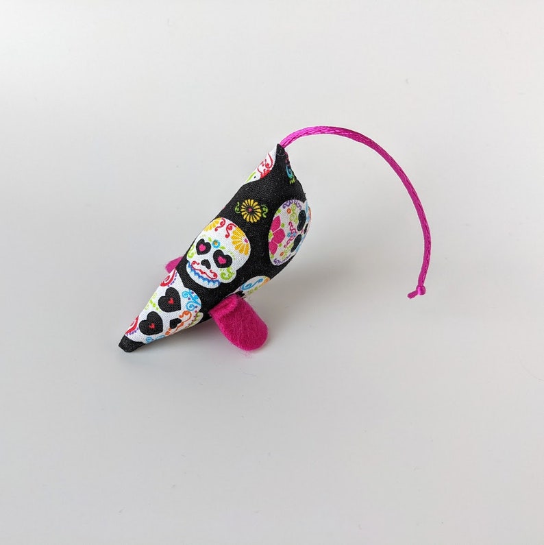May include: A black and white cat toy with a pink tail and pink ears. The toy is shaped like a mouse and has a sugar skull pattern.