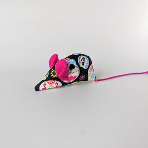 May include: A black and white cat toy mouse with a pink tail and pink ears. The mouse is covered in a fabric with a colorful sugar skull pattern.