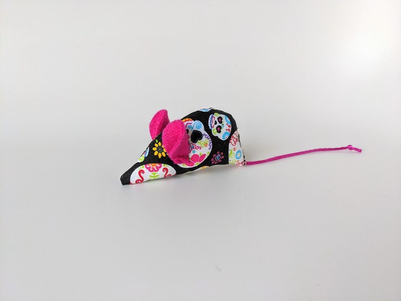 May include: A black and white cat toy mouse with a pink tail and pink ears. The mouse is covered in a fabric with a colorful sugar skull pattern.