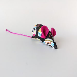 May include: A black and white fabric cat toy shaped like a mouse with pink ears. The fabric has a pattern of colorful sugar skulls.