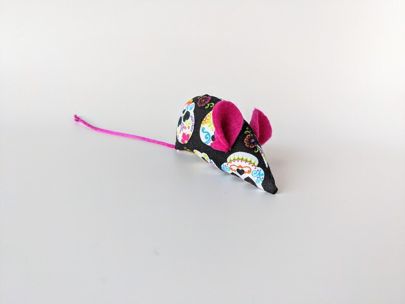 May include: A black and white fabric cat toy shaped like a mouse with pink ears. The fabric has a pattern of colorful sugar skulls.