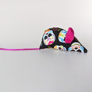 May include: A black and white cat toy shaped like a mouse with a pink ear. The mouse is covered in a fabric with a pattern of colorful sugar skulls.