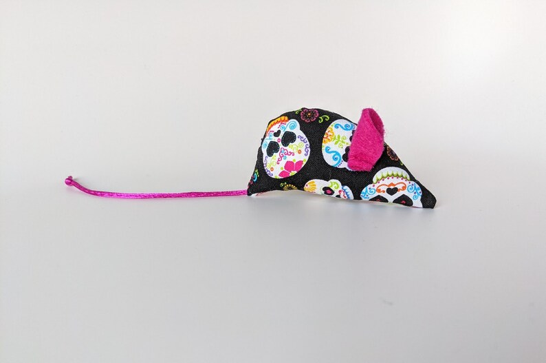 May include: A black and white cat toy shaped like a mouse with a pink ear. The mouse is covered in a fabric with a pattern of colorful sugar skulls.