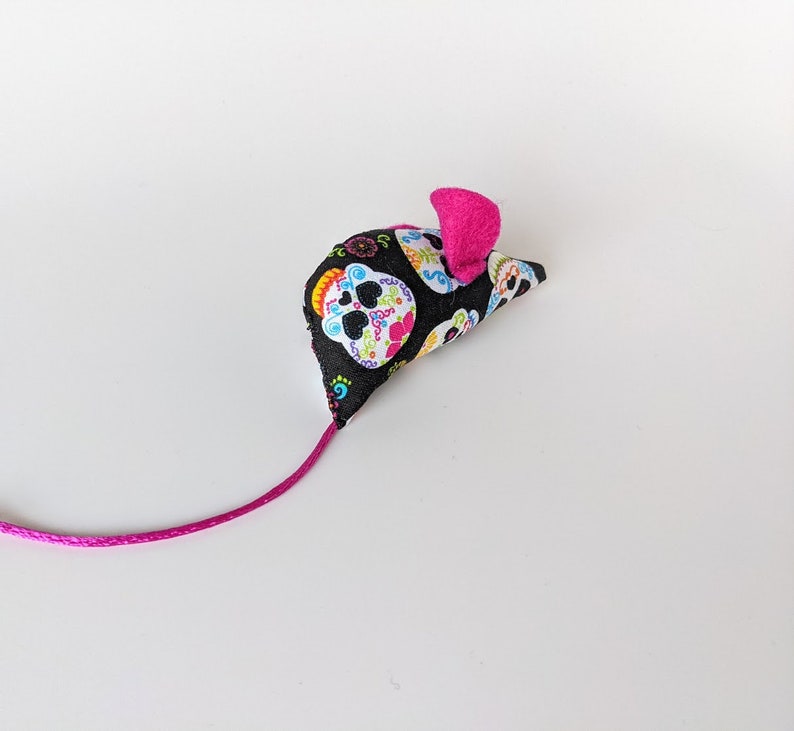 May include: A pink and black cat toy shaped like a mouse with a colorful sugar skull pattern.