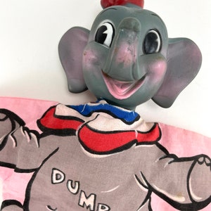 May include: A vintage hand puppet of Dumbo the elephant from Disney's animated film. The puppet is made of gray plastic with a pink face and red hat. The puppet is wearing a pink and blue costume with the word "DUMBO" printed on it.