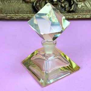 May include: A clear glass perfume bottle with a geometric design. The bottle has a square base and a diamond-shaped stopper. The edges of the bottle have a yellow tint, and the background is a light purple.