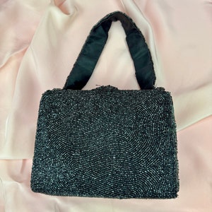 May include: A vintage black beaded handbag with a rectangular shape and a black satin handle. The bag's surface is covered in small, dark beads arranged in a swirling pattern. The background is a soft, pink fabric.