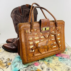 May include: A brown leather handbag with a woven design and a tooled leather bull on the front. The bag has two top handles and a zipper closure. The bag is sitting on a map of the United States.