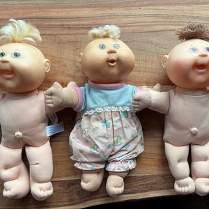 May include: Three dolls with round faces and soft bodies. The left doll has blonde hair, the middle doll has blonde hair and wears a light blue shirt with pink-patterned pants, and the right doll has brown hair. All have open mouths and pink cheeks.