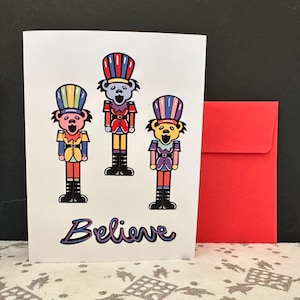 May include: A white greeting card with three cartoon bear figures dressed as toy soldiers, with the word "Believe" in script. A red envelope is next to the card. The bears wear colorful uniforms and hats.