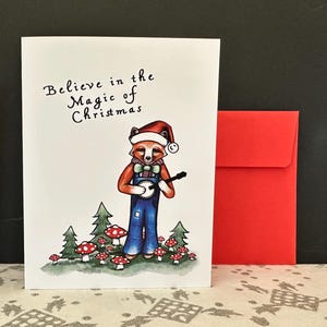 May include: A Christmas card with a whimsical illustration of a fox wearing a Santa hat and overalls, playing a banjo. The card reads "Believe in the Magic of Christmas." A red envelope is included.