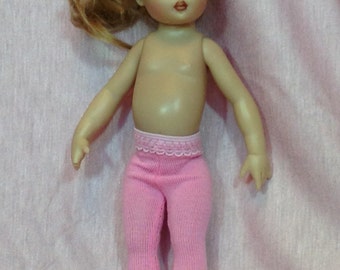 May include: A doll with light skin, blue eyes, and blonde hair tied in a side ponytail. The doll is wearing pink knit leggings that feature a delicate scalloped lace trim at the waistband. The doll has a neutral expression with painted red lips.
