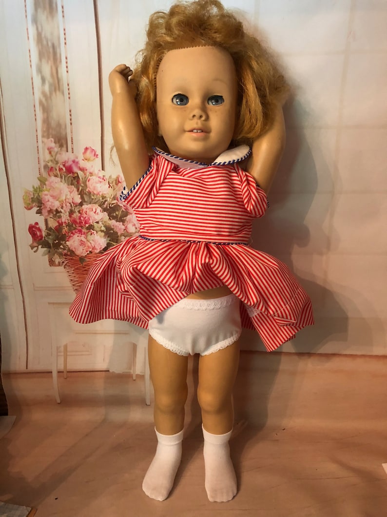 May include: A vintage red and white striped dress with a blue trim and white collar on a doll with blonde hair and blue eyes. The doll is wearing white socks and white underwear.