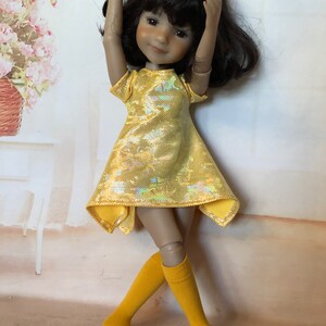 May include: A yellow dress with a shimmery iridescent finish and a high-low hemline. The dress is worn by a doll with brown hair and a bright smile. The doll is wearing yellow knee-high socks.