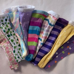 May include: A collection of ten colorful fabric doll leggings with white lace trim. The leggings feature various patterns including polka dots, stripes, and floral designs.