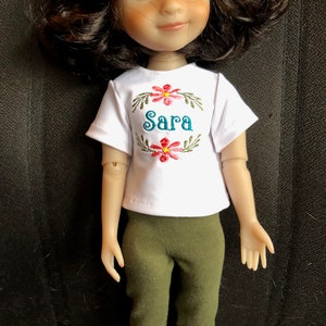 Puede incluir: Una camiseta blanca con un diseño floral verde y el nombre "Sara" bordado en la parte delantera. La muñeca lleva pantalones capri verdes.