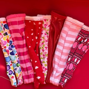 Può includere: Un set di otto paia di leggings per bambole con motivi rosa e rossi. I leggings hanno diversi motivi, tra cui cuori, strisce e disegni geometrici.