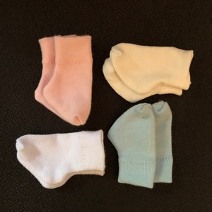 Puede incluir: Cuatro pares de calcetines de diferentes colores: rosa, blanco, crema y azul claro. Los calcetines están doblados y dispuestos en forma de cuadrado sobre un fondo oscuro.