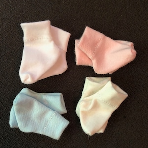 May include: Four pairs of baby socks in white, pink, light blue, and a cream color. The socks are folded and arranged on a black surface.