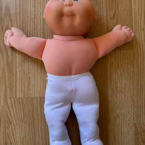 May include: A vintage baby doll with a peach-colored body and a round face. The doll has blue eyes, a small tuft of blonde hair, and is wearing white pants. The doll's arms are outstretched, and it has a smiling expression.