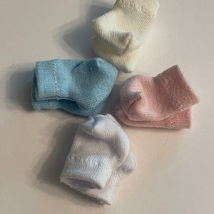 May include: Four pairs of baby socks in pastel colors: white, light blue, and pink. The socks are made of a soft, knitted material and are arranged on a white surface. The socks are small and designed for infants.