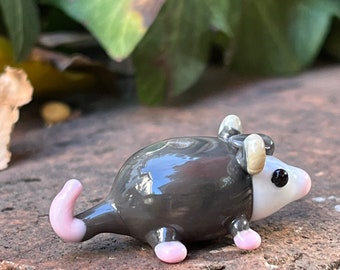 May include: A handcrafted glass opossum figurine. The figurine features a gray body, white face, and pink feet and tail. The opossum has small, rounded ears and black eyes. The glass is glossy and reflects light.